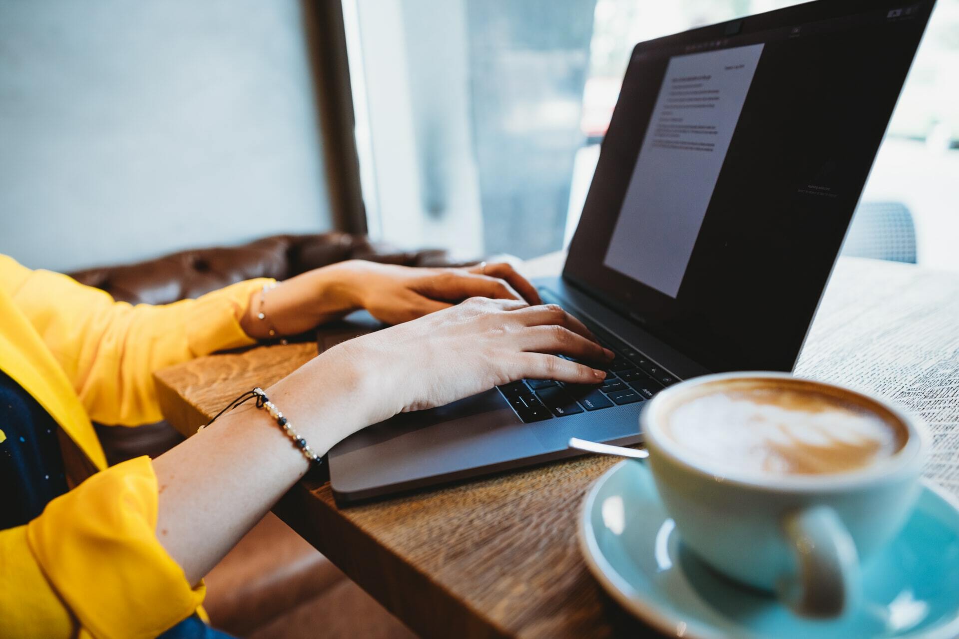 A laptop with a person's hands on the keyboard and an espresso-based drink next to the laptop